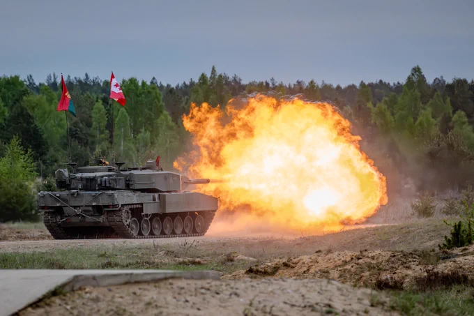 A tank fires its main gun, producing a large fireball and dust cloud on a sandy training range adorning a Canadian flag and a regimental flag.