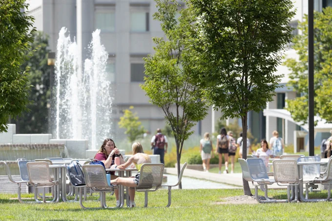 People are sitting at outdoor tables on Missouri State's campus with Strong Hall in the background. A fountain is visible, and trees provide shade. Students are walking in the distance.