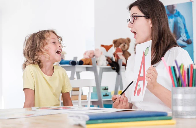 A person is holding a printed sheet with a large letter "A" and cherries, engaging with a young individual who is smiling. In the background, stuffed animals and art supplies are visible on shelves.