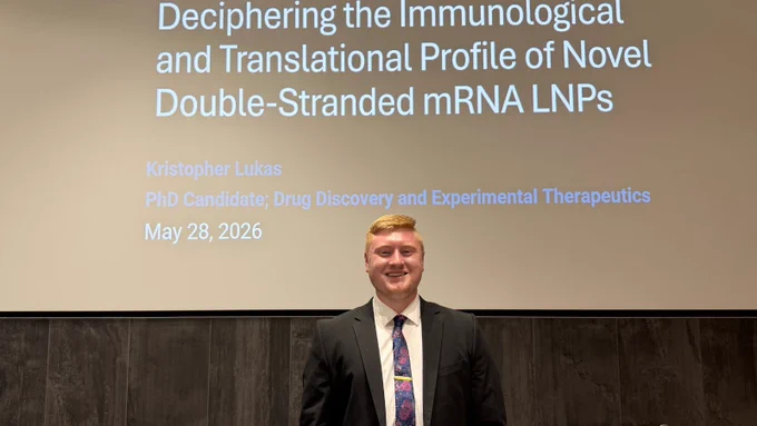 Photo of a PhD candidate Kris Lukas standing in front of a presentation screen after a dissertation defense. The slide reads, “Deciphering the Immunological and Translational Profile of Novel Double-Stranded mRNA LNPs,” and identifies Kristopher Lukas, PhD Candidate in Drug Discovery and Experimental Therapeutics, with a defense date of May 28, 2026. He is wearing a dark suit and patterned tie and smiling toward the camera.
