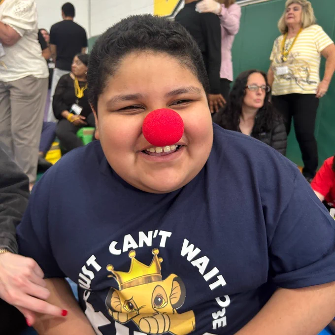 Child wearing a navy blue t-shirt with a crowned lion graphic and the text 'Just can't wait to be king' sitting indoors among a group of people.