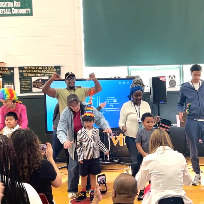 Children and adults performing on a gym stage with colorful costumes and hats, while an audience captures the moment on phones.