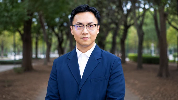 Individual wearing glasses and a blue suit standing outdoors on a tree-lined pathway.