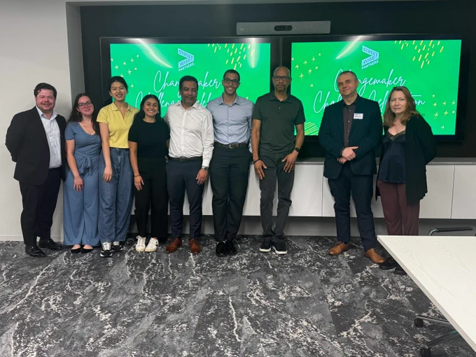Group of nine professionals standing in front of two green screens displaying 'Changemaker' text in a modern office setting.