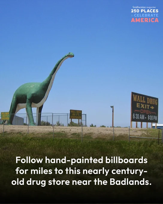 A dinosaur statue stands beside signs for Wall Drug, set against a clear blue sky. A fence is visible in the foreground, with grassy terrain surrounding the displays.