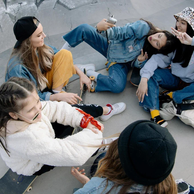 A group of people sitting in a circle at a skate park, taking a selfie with a smartphone. They are wearing casual clothing, including denim jackets, hoodies, and caps. Skateboards and backpacks are visible near them.