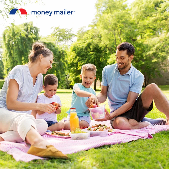 Family of four enjoying a picnic on a pink blanket in a sunny park surrounded by green trees.