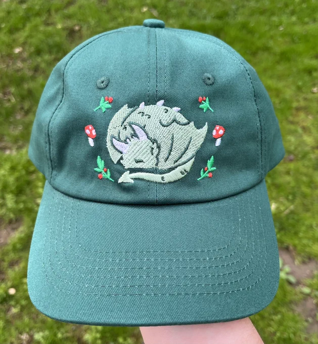A teal hat featuring an embroidered dragon design. The dragon is sleeping with its tail curled next to its face. It is surrounded by little plants and mushrooms.