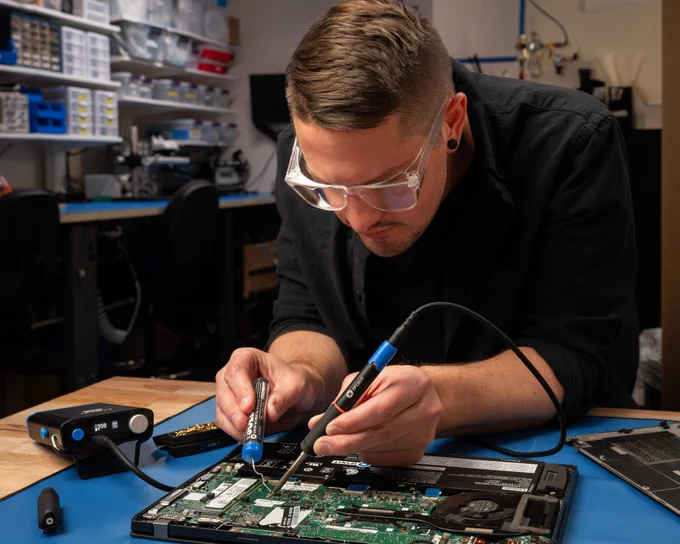 A person wearing clear safety glasses uses a soldering iron and solder wire to repair a laptop motherboard at a workbench. An iFixit FixHub portable soldering station sits nearby, with electronic components, tools, and a partially disassembled laptop visible in a workshop setting.