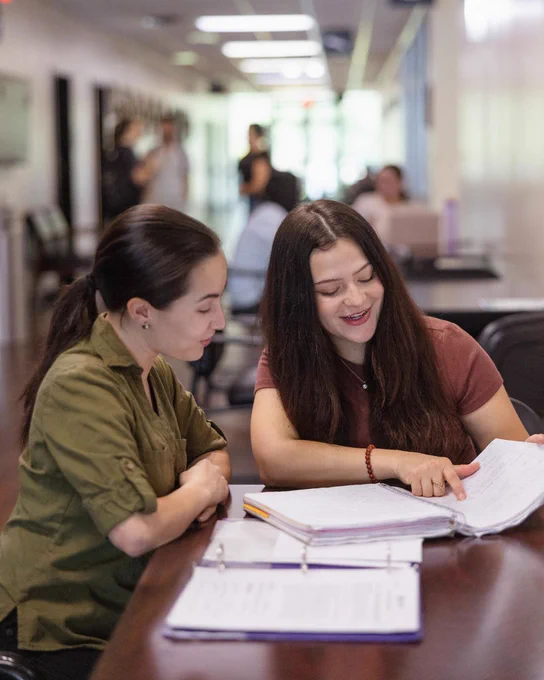 Two Southeastern students sit together at a table in a campus hallway, reviewing notes and coursework in an open binder. One student points to a page while they collaborate and smile, with other students visible in the background.