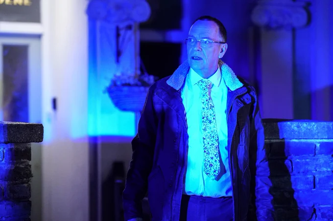 EastEnders' Ian Beale stands outside his house, illuminated by blue-tinted police lights. He is wearing glasses, a patterned tie, and a jacket with a furry collar. 