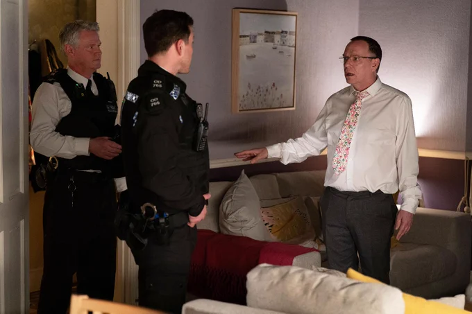 Two uniformed police officers stand in Ian Beale's living room in EastEnders, engaging in conversation with him. He's wearing a white shirt and colorful tie. A framed artwork hangs on the wall above a beige couch with decorative cushions.
