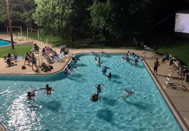 People enjoying a nighttime swim while watching a movie in a well-lit outdoor pool surrounded by trees and seating areas.