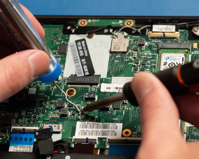 Close-up view of a laptop motherboard during a soldering repair. A soldering iron and solder wire are being applied to a small surface-mounted electronic component on the circuit board, demonstrating precision electronics repair and microsoldering work.