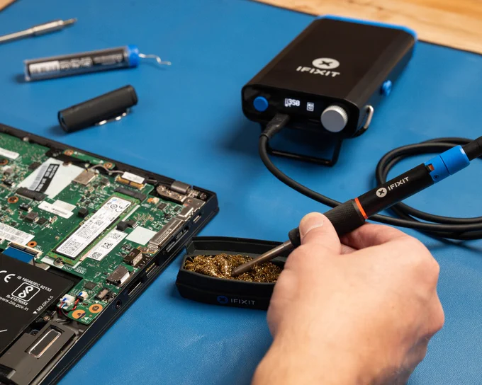 A hand holds an iFixit soldering iron above a brass tip cleaner while working on an open laptop. In the background, an iFixit FixHub portable soldering station powers the iron, with soldering tools and exposed computer components arranged on a blue work mat. The image highlights electronics repair and soldering workstation setup.