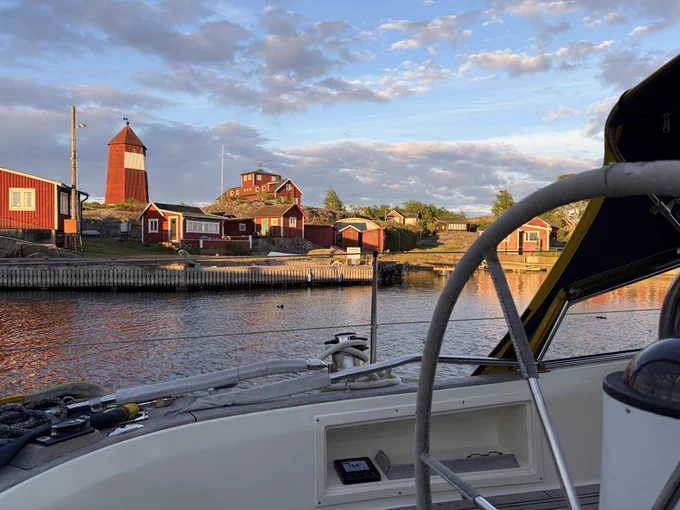 Abendstimmung in einem Hafen in den Schwedischen Schären, von einem Segelboot aus fotografiert. Man sieht im Vordergrund Teile des Cockpits und das Steuerrad. In der Bildmitte eine Wasserfläche mit Vögeln. Darüber eine holzverkleidete Kaimauer und darüber ein paar einfache rote Holzhäuser. Über den Häusern ist ein rot weißer Holzturm, der alte Lotsenturm. Darüber ein rötlicher Abendhimmel mit ein paar Wolken.
