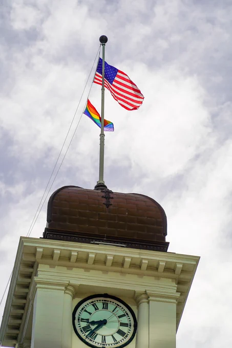 Old City Hall with an American flag and a rainbow flag flying at the top against a cloudy sky.