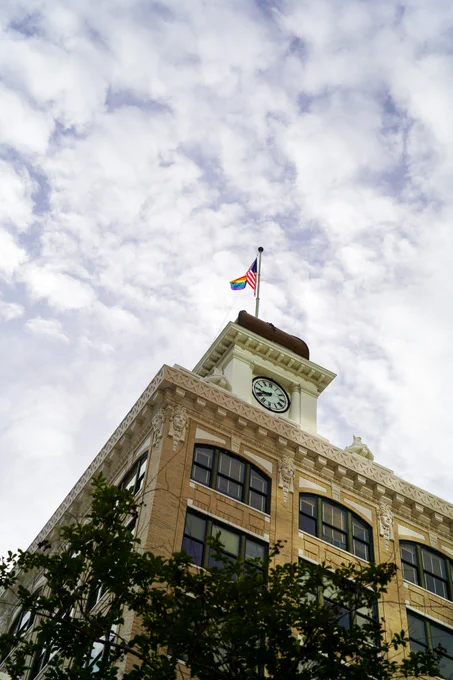 Old City Hall with some trees in the foreground, with an American flag and a rainbow flag flying at the top against a cloudy sky.