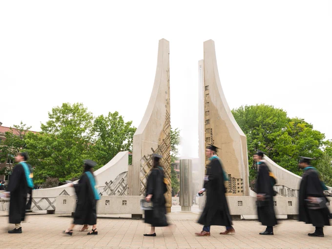 Graduates in caps and gowns walk past the Purdue University Gateway to the Future arch, with trees and university buildings in the background.