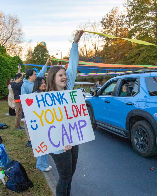 A volunteer holding a sign that says “honk if you love Camp”, while they are welcoming cars into cabin row.