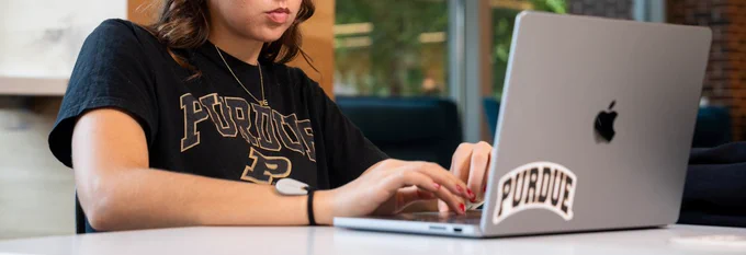 Woman wearing black shirt with Purdue on it is sitting down and typing on a silver computer with a Purdue sticker on it. 