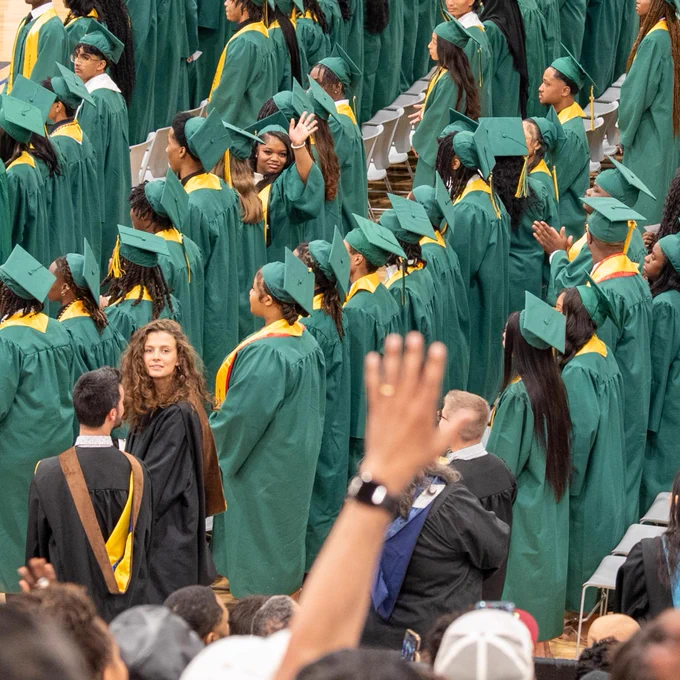 A large group of graduates wearing green caps and gowns at a commencement ceremony, some raising their hands.