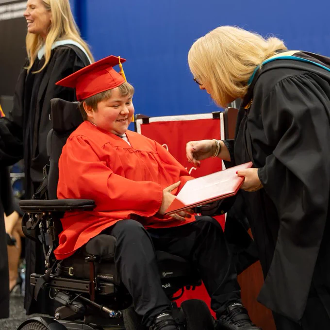 A young person in a red graduation gown and cap receiving a diploma from an adult in a black graduation gown.