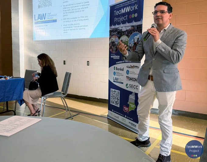 Speaker stands at the front of a community room holding a microphone and presenting on open work permits, with a projected LAW (Legal Assistance of Windsor) slide behind and a TeaMWork Project banner beside them; a seated attendee is visible at a nearby table.