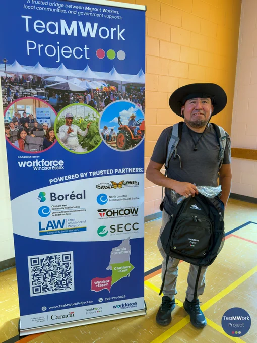 	Un participante en el taller posa junto a una pancarta del proyecto TeaMWork con una mochila que ha recibido en el evento, en el mismo espacio comunitario donde se celebró la sesión.
