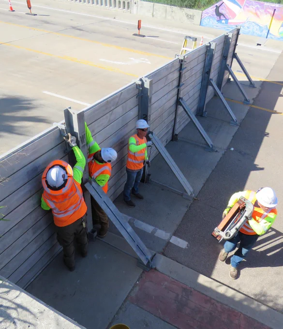 Construction workers in orange safety vests and helmets securing a temporary wooden barrier along a roadside.