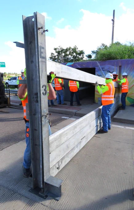 Construction workers in orange safety vests installing a large metal floodgate on a concrete surface outdoors.