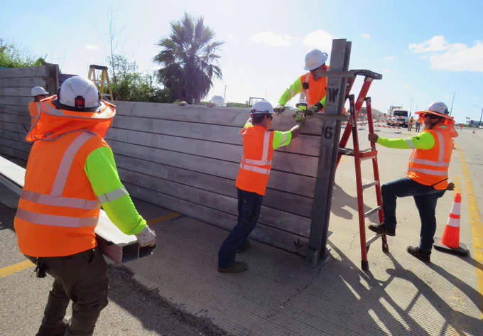 Four construction workers in safety vests and helmets assembling a large concrete barrier on a sunny road.