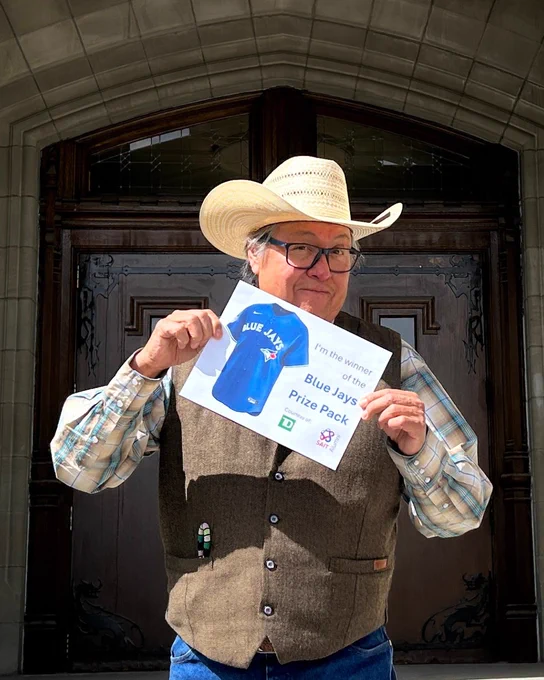 A person in a cowboy hat and vest holds a Blue Jays prize pack certificate in front of a large wooden door.