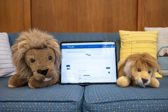The lions Sol and Edgar are sitting on a blue couch at the State Library. In between them is an iPad and on the screen is the SC Department of Insurance digital collection.