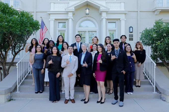 A group of people, all dressed in formal attire, stand on the steps of a building with columns. Some are giving a thumbs-up gesture. UTEP President Heather Wilson stands in the center of the front row. An American flag is visible to the side.
