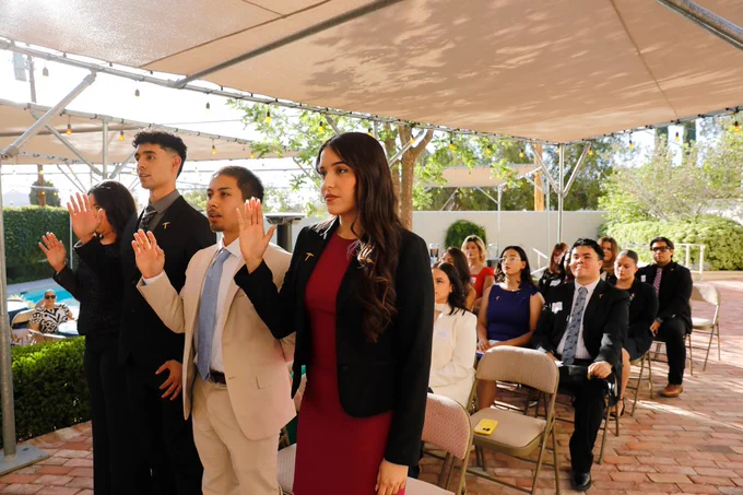 A group of people seated outdoors, raising their right hands in an oath-taking gesture. The setting includes a brick floor, chairs, and a shaded canopy. In the background, a pool and lush greenery are visible.