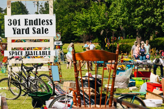 Outdoor yard sale with a large sign reading "301 Endless Yard Sale" and "Space Available" in the foreground. Bicycles, a wooden chair, and assorted household items are displayed on the grass, while several people browse tables filled with colorful goods. Trees and greenery are visible in the background.