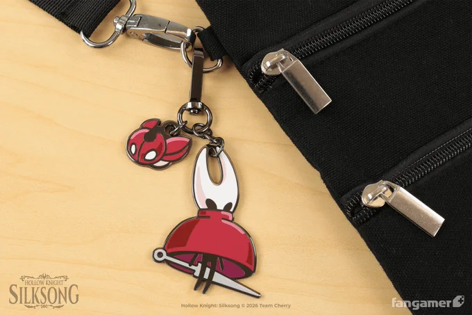 A photo of the Drifting Hornet keychain, recently released in our Hollow Knight: Silksong collection, hanging from a black bag. It's a double-sided, hard-enamel keychain charm showing Hornet floating in the Drifter's Cloak, paired with a single-sided hard-enamel Cogfly charm. 