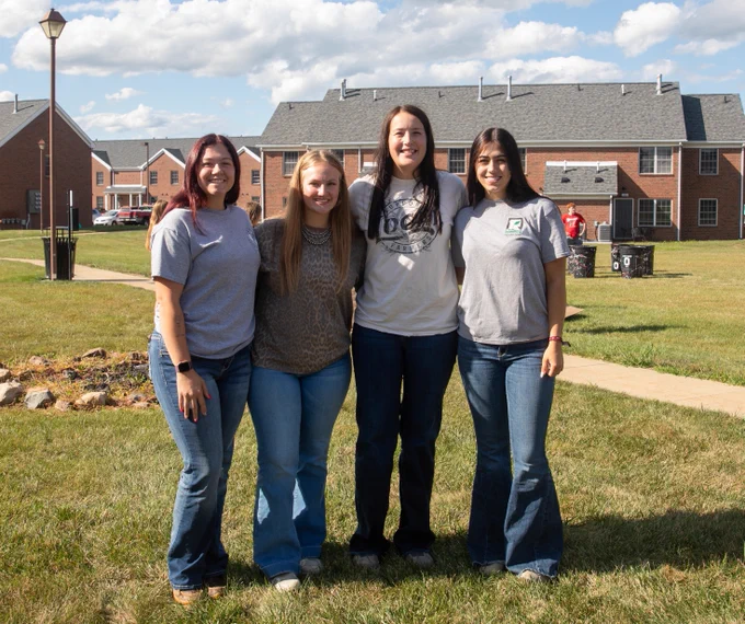 Four people are standing together on a grassy area with brick buildings in the background. They are smiling at the camera, wearing casual clothing, and the sky is partly cloudy.