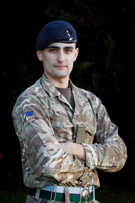 A British soldier is standing, arms folded, facing the camera. He is wearing  a camouflage uniform, and a blue beret on his head. 