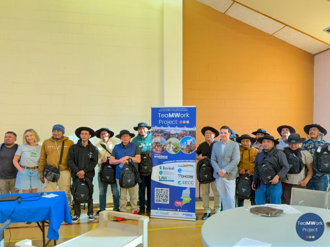Group of workshop participants stand in a line indoors holding TeaMWork backpacks, posing for a photo in front of a TeaMWork Project banner following the legal education session.