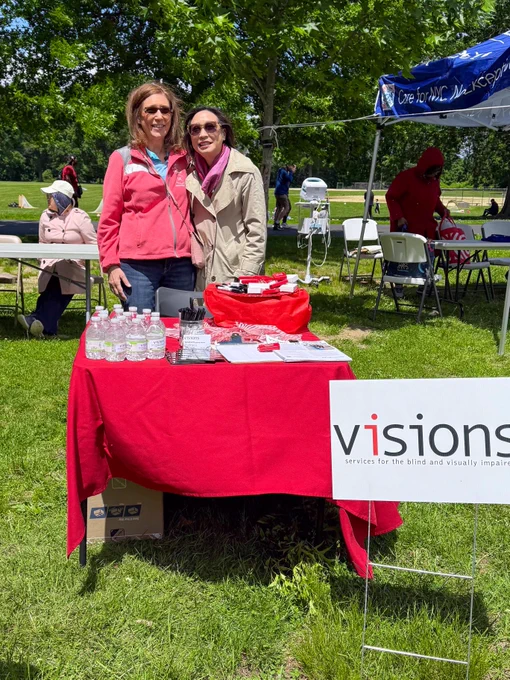 Outside in a park, Doreen DiLeonardo (left) stands next to Councilwoman Linda Lee, as they both stand behind a red table decorated with VISIONS information and promotional items and signage. 