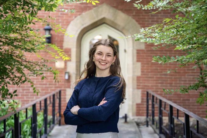 Ella Fahey stands with arms crossed and smiling in front of a gothic stone archway on Duquesne University's campus, framed by green trees.