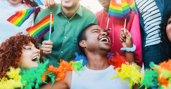 A group of people celebrating with joy, waving rainbow flags. One person is draped in colorful feather boas, smiling widely. The scene is vibrant and filled with cheerful expressions, representing a festive atmosphere.