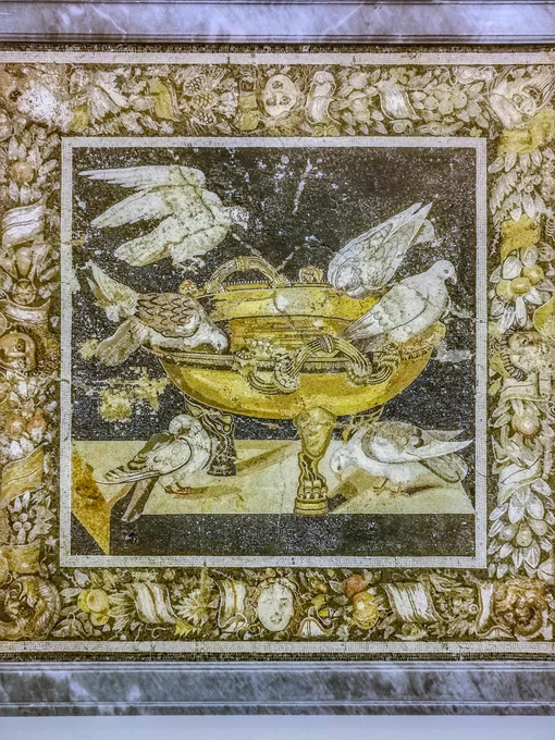 MOSAIC EMBLEMA WITH DOVES, 120-100 BCE, FROM THE HOUSE OF THE GEOMETRIC MOSAICS, POMPEII, NOW IN THE MUSEO ARCHEOLOGICO NAZIONALE DI NAPOLI (MANN, INV. 114281)

Crafted in microscopic opus vermiculatum by Greek-trained masters in Campania, this magnificent mosaic emblema depicts doves at a bronze cantharus. This variant of the "Pliny's Doves" mosaic includes two extra doves on the ledge below, while an upper bird is caught in an almost photographic snapshot, landing with wings outspread. The composition shows the matte nature of local stones (dark limestones and dense volcanic deposits), punctuated by vibrant glass-paste highlights rendering the wet sheen of water and glinting plumage. Framed by a rich border of theatrical masks and fruit garlands, this Pompeian original prioritises a tactile, muscular naturalism, differing sharply from the later imperial Capitoline Museums version's ethereal, highly polished golden basin and smooth painterly illusionism. 