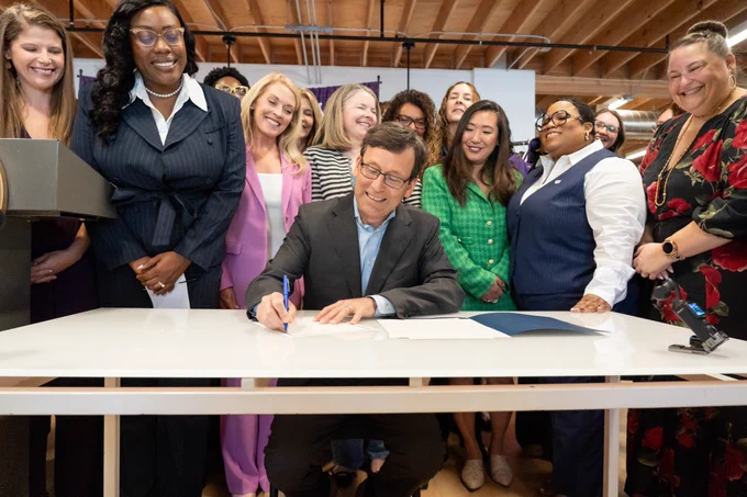 Bob seated at table signing executive order with large group of individuals smiling behind him 