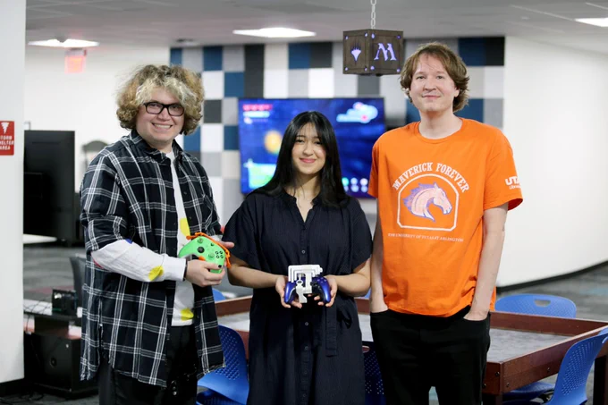 Pictured is a photo of two a UTA Libraries staff member, student worker and a TWU student. In the photo they are holding two video game controllers that have been adapted with snap-on modification so that they can be used by everyone.