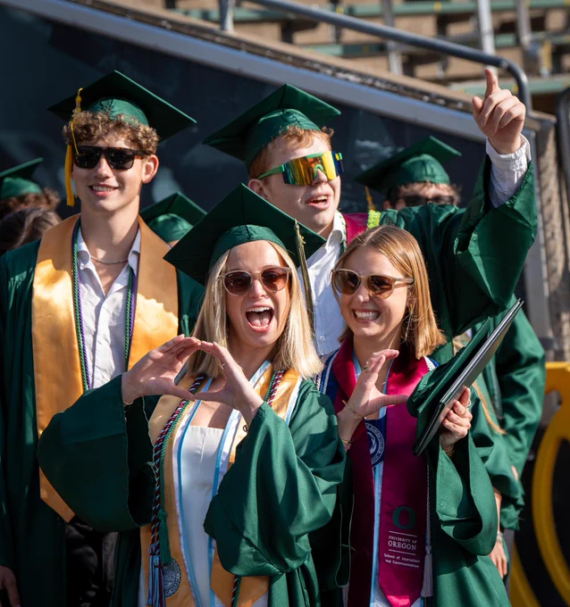 A group of graduates in green caps and gowns celebrate together. One is throwing the O with their hands. Another is pointing upwards, while others smile. A "University of Oregon" sash is visible. The background shows stadium seating.