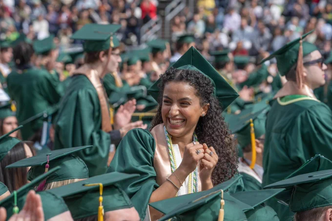Graduates in green caps and gowns are seated outdoors at a commencement ceremony. One individual in the foreground is smiling broadly and clasping their hands. The atmosphere appears festive and celebratory.