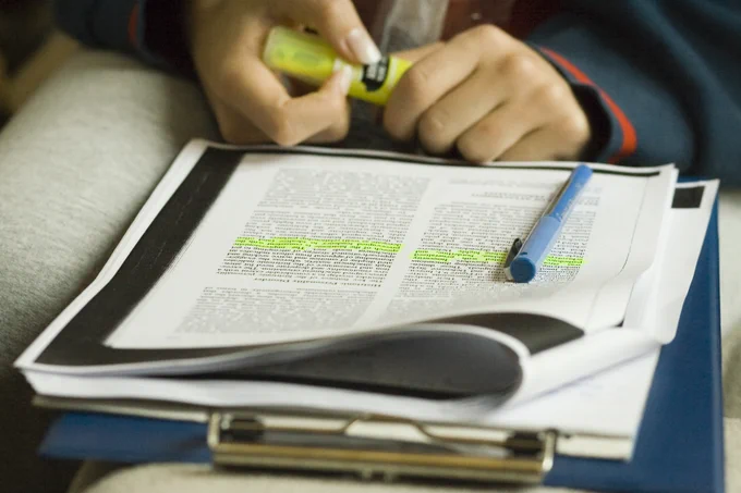 A person is highlighting a passage on a printed document with a yellow marker. A blue pen rests on the paper, and the document is placed on a clipboard.